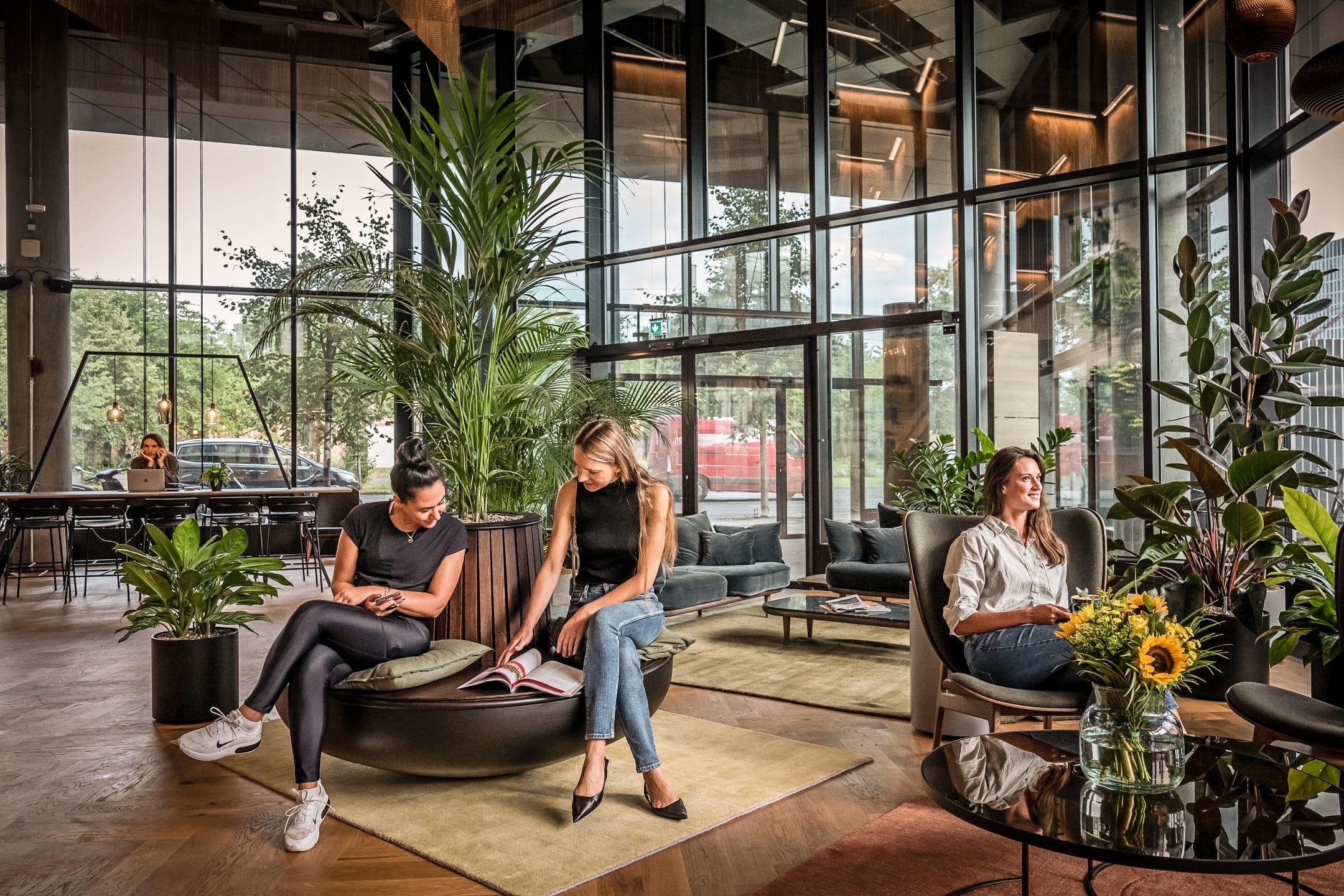 A modern office lobby featuring large windows, lush green plants, and contemporary flexible workspace solutions. Three people, one sitting and two talking, create a relaxed, engaging atmosphere. sunflowers and area rugs add An image from EDGE Workspaces.