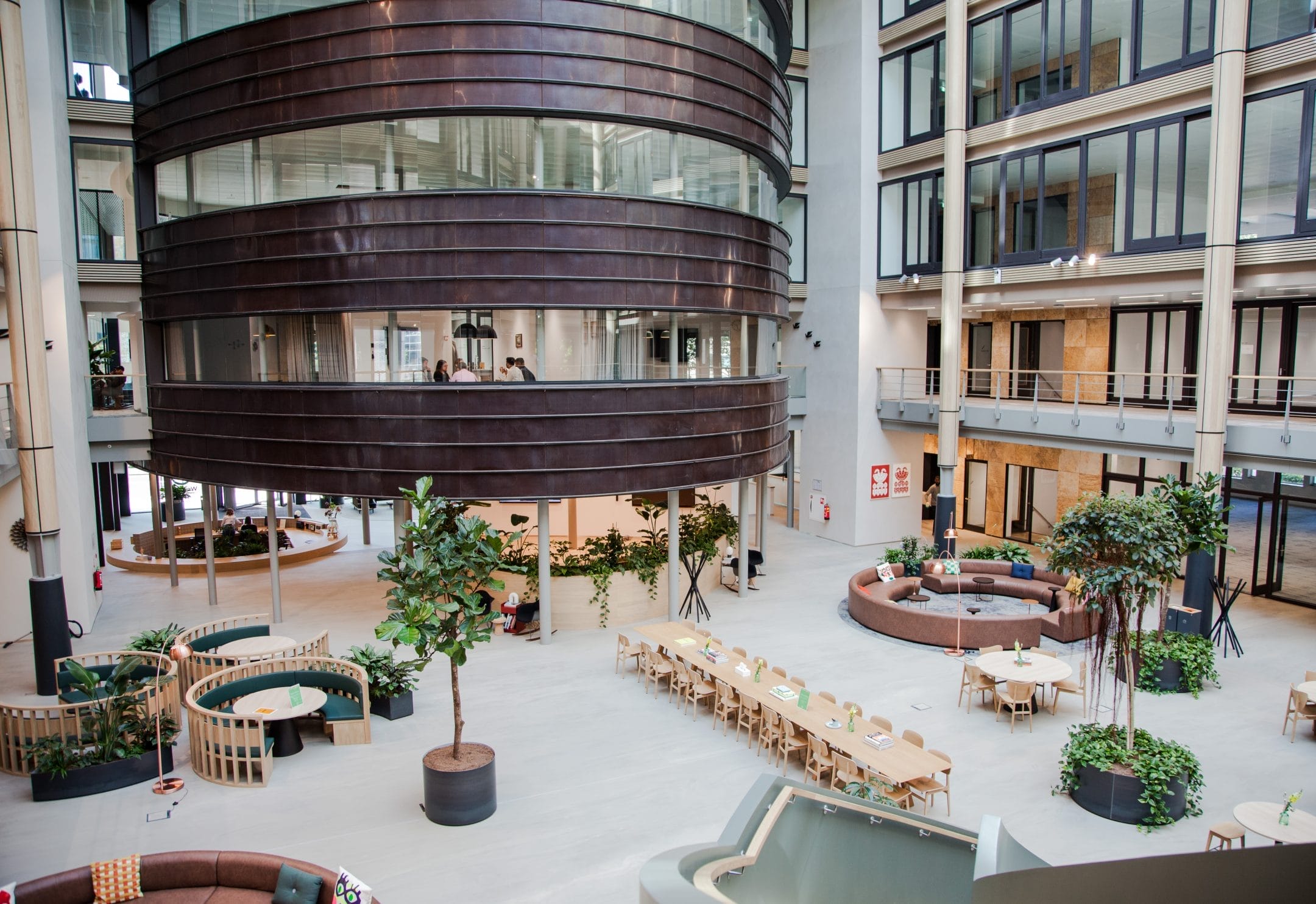 Modern coworking office atrium with a circular wooden structure in the center, surrounded by green potted plants, casual seating areas, and multi-level balconies with glass railings. An image from EDGE Workspaces.