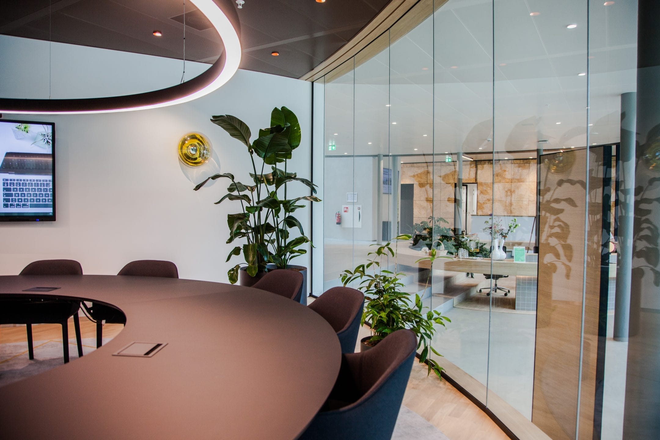 Modern office meeting room with a round dark table, surrounded by chairs. The room features glass walls, a large potted plant, and a mounted digital screen displaying a calendar. Decorative elements include a An image from EDGE Workspaces.