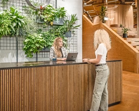 Ein moderner Empfangsbereich in einem Büro, in dem zwei Frauen miteinander kommunizieren. Eine mit lockigem blondem Haar arbeitet an einem Laptop hinter einer mit grünen Pflanzen geschmückten Holztheke, während die andere ihr gegenübersteht und in ein Gespräch vertieft ist. An image from EDGE Workspaces.