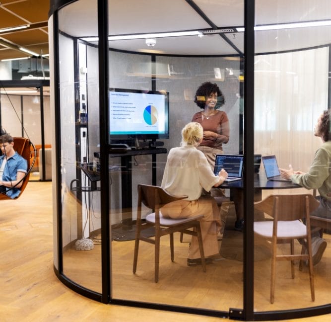 A diverse group of professionals participates in a meeting inside a modern glass-walled office pod, an innovative coworking environment. A woman stands presenting a pie chart on a screen while others listen attentively, An image from EDGE Workspaces.